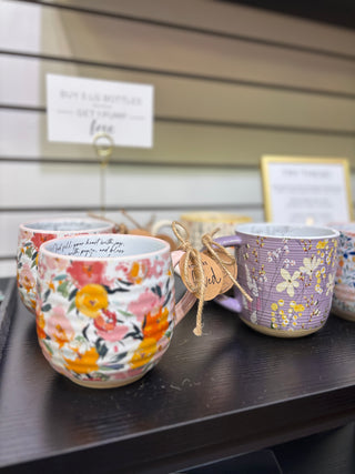 Two decorative mugs on display, one floral with pink handle and “You are loved” charm tag