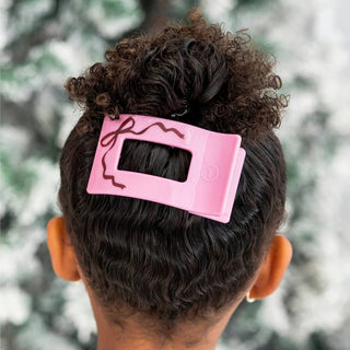 Pink flat rectangle hair clip with bow graphic, clipped on dark curly hair