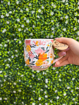 Hand holding floral mug with pink handle and “You are loved” charm tag outdoors