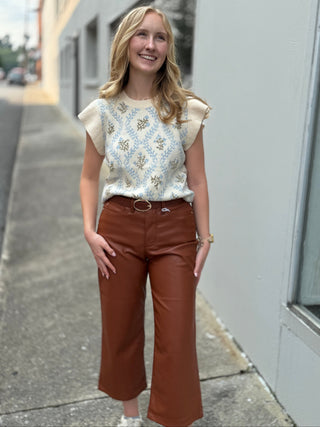 A woman stands on an outdoor sidewalk wearing a cream and blue patterned short-sleeve knit top with brown wide-leg cropped pants and white sneakers.